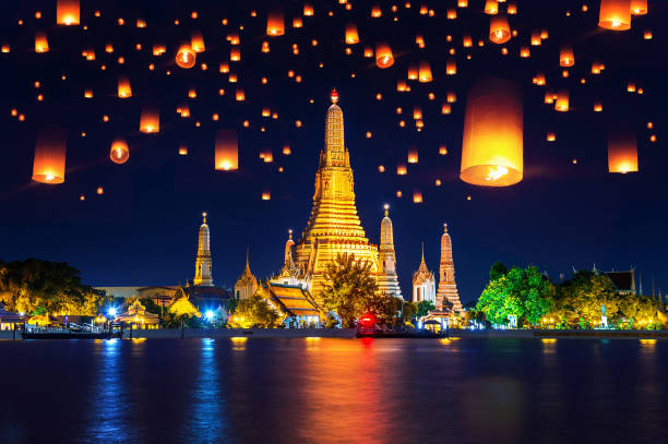 Glowing lanterns floating in the night sky by a river and golden temple during Loy Krathong Festival in Thailand